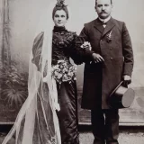Ein Brautpaar von 1902 Ein Brautpaar steht eingehenkelt, mit etwas Abstand zueinander. Die Braut trägt ein schwarzes Kleid. 1902
